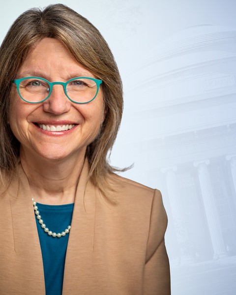 Sally Kornbluth wearing glasses and tan jacket in front of blue graphic of MIT Dome