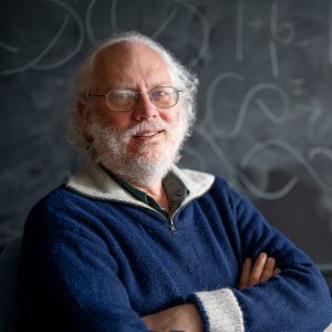male with glasses and white hair and beard standing in front of chalkboard with physics equations