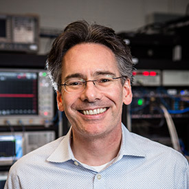 male with glasses and dark hair wearing pale blue collared shirt standing in front of computing equipment