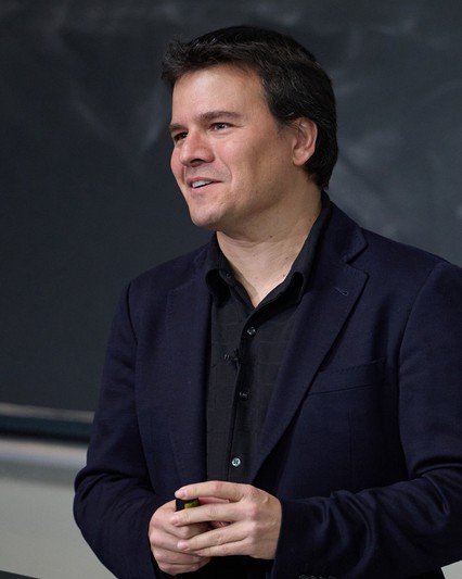 male standing in front of blackboard wearing dark shirt and blue jacket