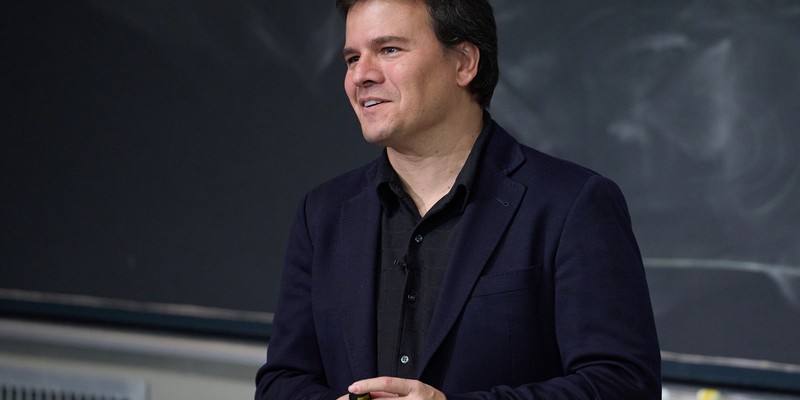 male standing in front of blackboard wearing dark shirt and blue jacket