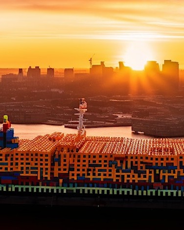 large container ship with stacks of hundreds of orange shipping containers docking into city port at sunset