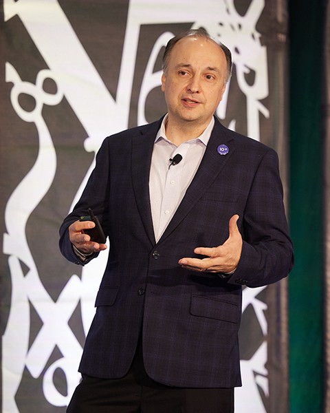 Large white numbers and mathematical symbols appear behind male speaker wearing dark suit and white shirt.