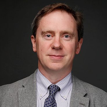 headshot of middle-aged male wearing grey suit, light blue shirt with dark blue tie in front of dark grey background