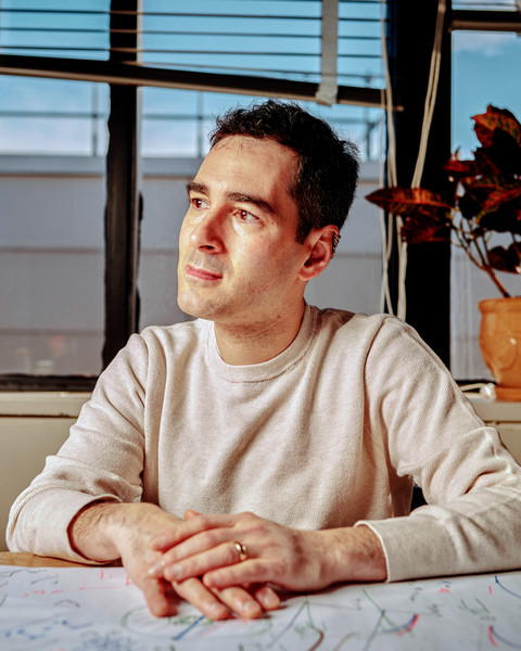 Rafael Jaramillo sits in his office and looks to the side. A large wrench sits on the window sill. The desk is covered in white paper with many drawings and notes on it.