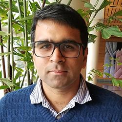 man wearing glasses, white collared shirt and blue sweater in front of leafy green plants