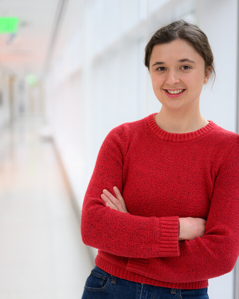 Miranda Schwacke wearing a red sweater and standing in a brightly lit hallway