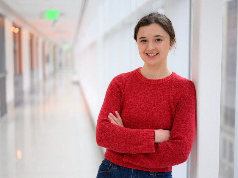 Miranda Schwacke wearing a red sweater and standing in a brightly lit hallway