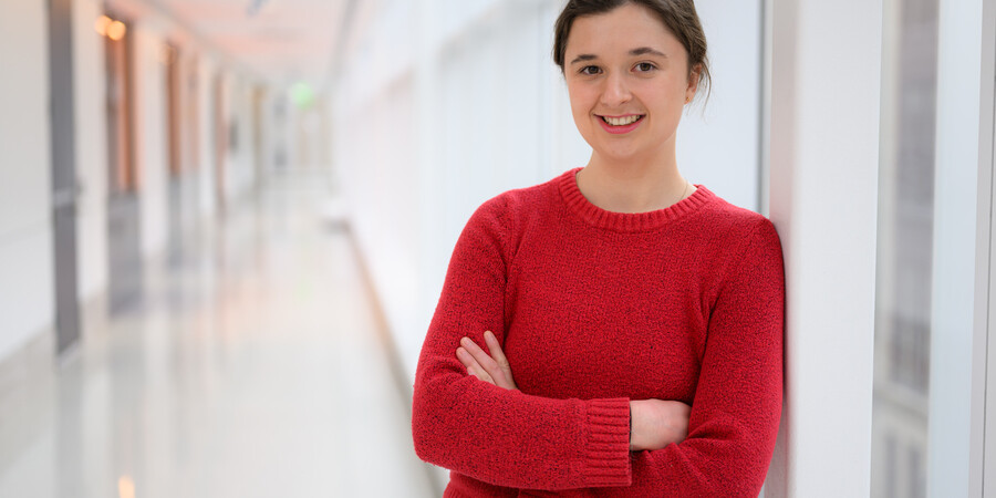 Miranda Schwacke wearing a red sweater and standing in a brightly lit hallway